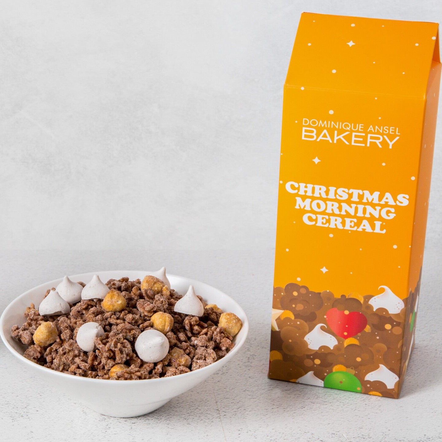 A bowl of Christmas Morning Cereal with milk and a packaged cereal box with 'Christmas Morning Cereal' text and festive design in the background.